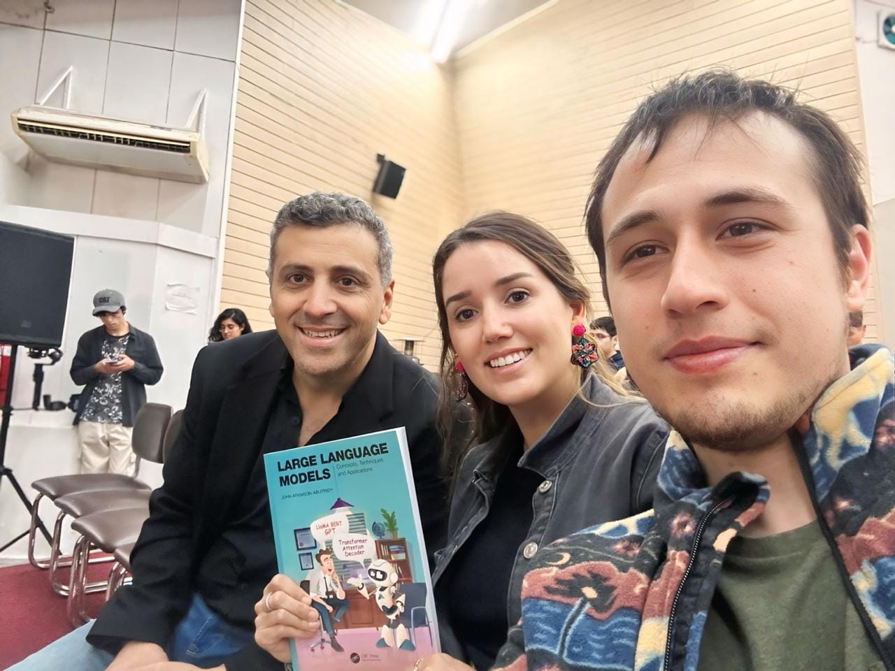 Foto de John Atkinson-Abutridy, Fernanda Herreros y Diego Orrego (de izquierda a derecha). Fernanda sostiene su premio, el libro 'Large Language Models', ganado durante la presentación de John.